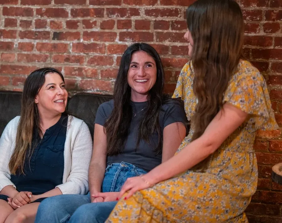 Three women talking on a couch at a Hoboken Grace dinner group, one sitting on the arm beside them